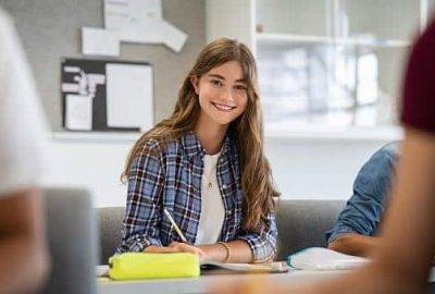 Beautiful university student smiling while studying for exams in classroom. Pretty woman sitting in classroom full of students during class. Portrait of happy young woman writing notes and looking at camera.